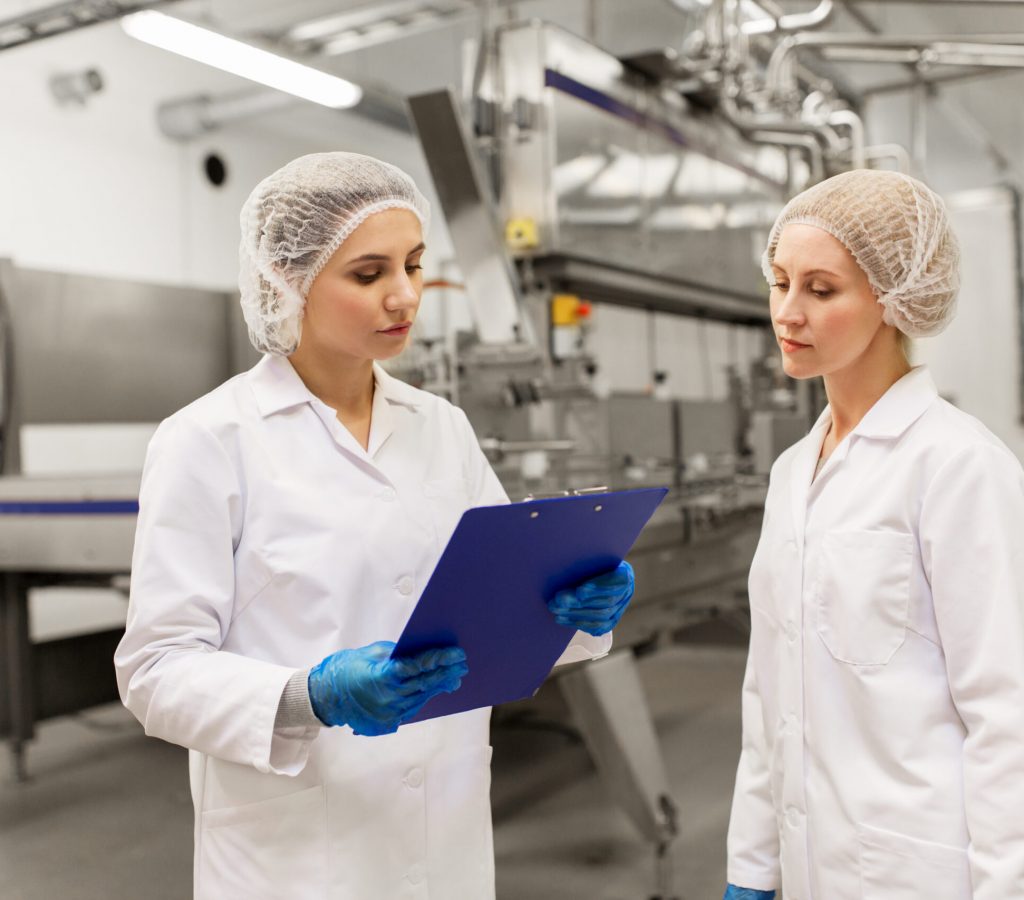 manufacture, industry and people concept - women technologists with clipboard at ice cream factory shop